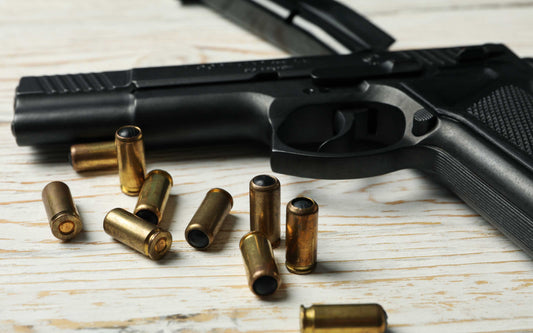 Black semi-automatic handgun on a light wooden surface surrounded by brass-colored bullets, some upright and some lying flat.