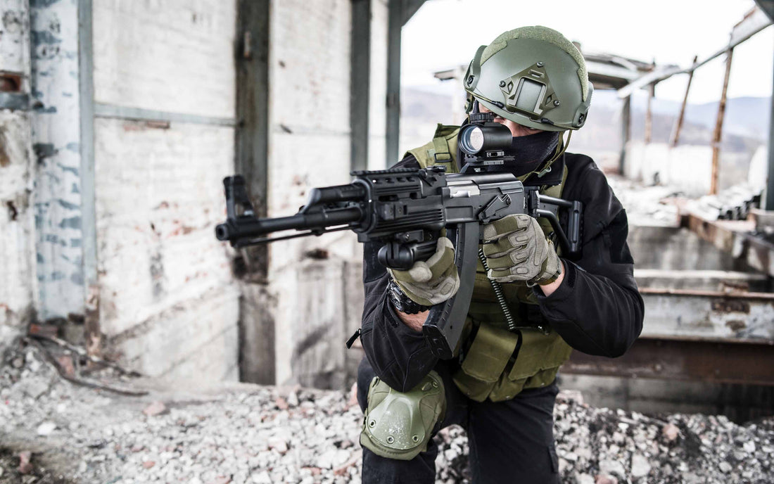 Person in tactical military gear aiming a scoped rifle inside a damaged industrial building with broken concrete and exposed steel beams