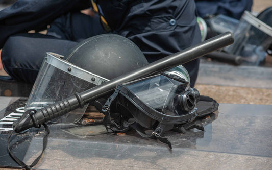 Close-up of riot control gear on the ground, including a black helmet with face shield, a baton, and a gas mask, with uniformed personnel standing nearby