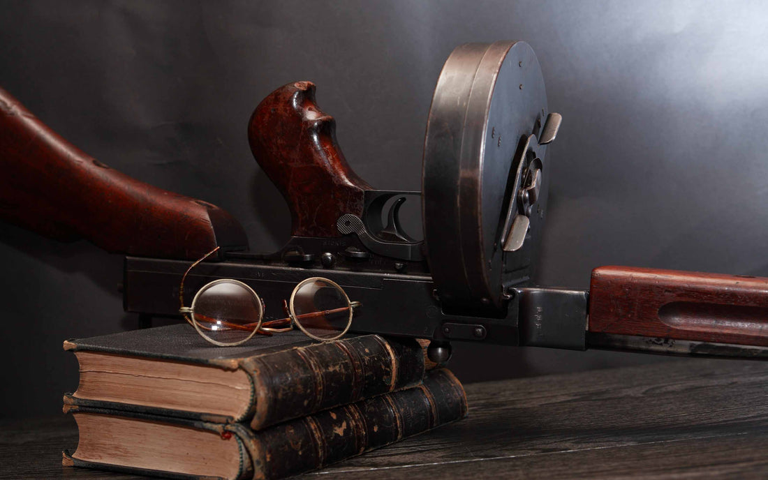 A vintage firearm with a wooden stock and drum magazine resting on stacked old books, with round eyeglasses in front