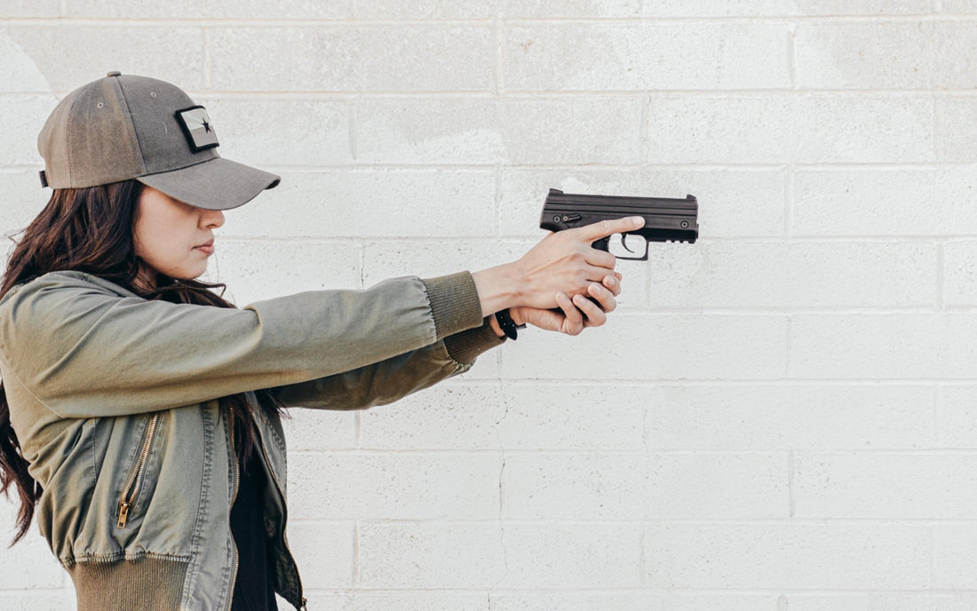 A person wearing a green jacket and a cap is aiming a black handgun at an unseen target against a white brick wall.
