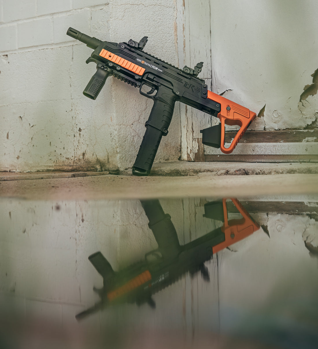 Left side byrna TCR rifle with reflection in a puddle leaning on a white concrete wall. 