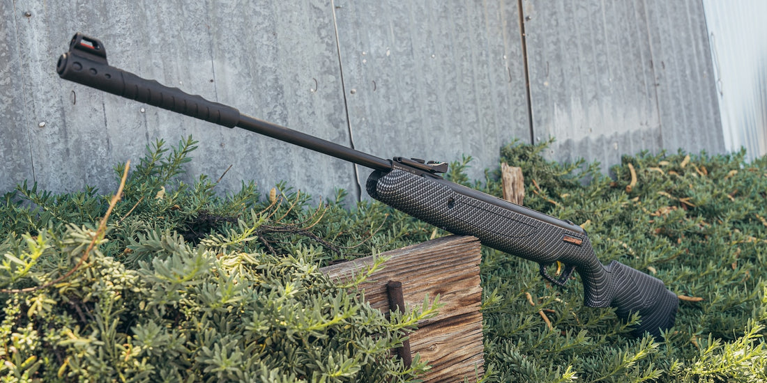 Left side of Ceonic air rifle leaning on a piece of wood in grass, with a grey fence in the background. 